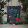 Colorful tapestry with jungle design held by a person against a white paneled wall with green plants.