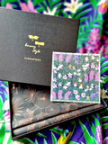 Close-up of an open black Binary Style gift box with a floral design postcard inside, on a colourful floral background